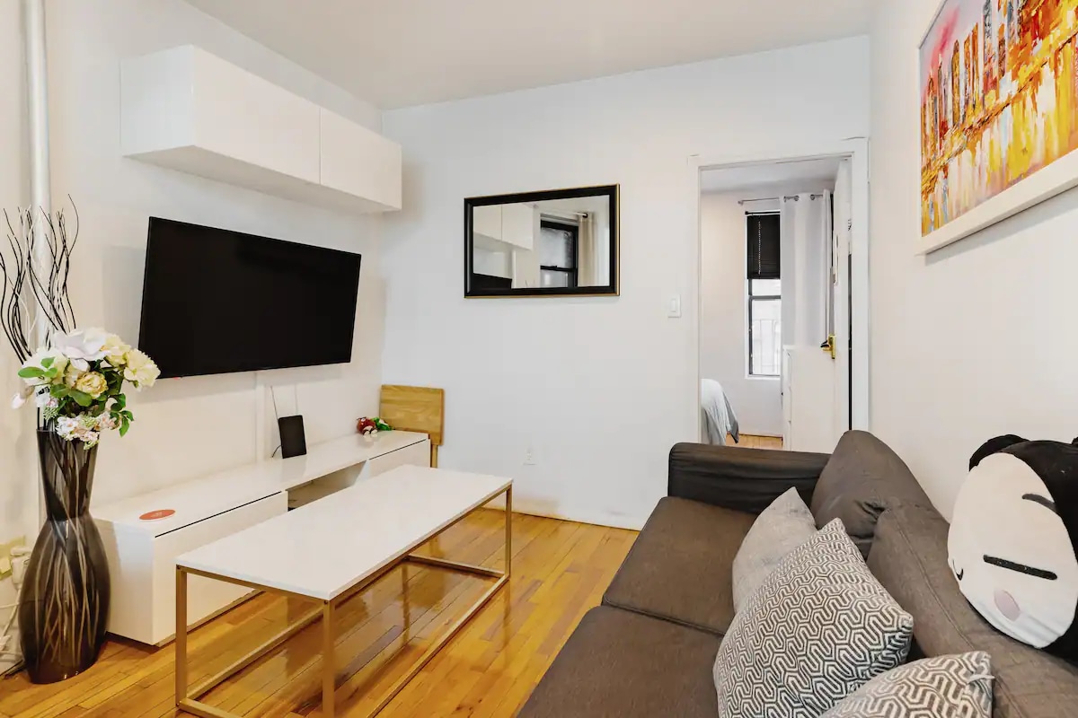 A modern living room showcases a sleek flat-screen TV mounted on a white wall. A light wood coffee table is centered in front of a comfortable sofa with decorative cushions. A simple chair and a mirror reflect the space's brightness, while a colorful artwork adds a touch of vibrancy.