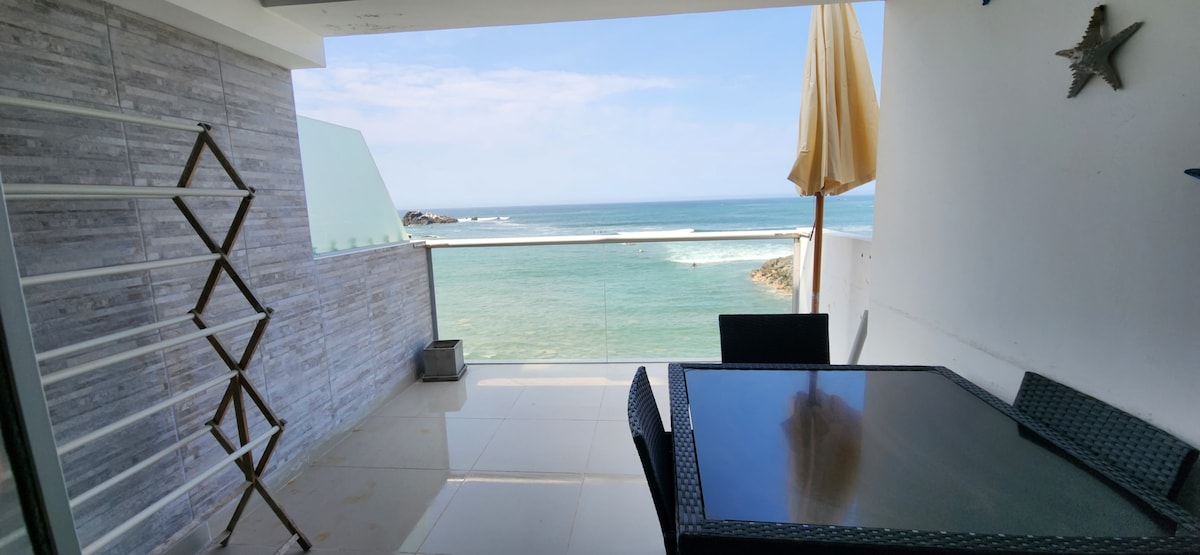 A balcony area is visible, featuring a dark outdoor dining table surrounded by matching chairs. The ocean can be seen in the background, with waves gently lapping at the shore. A sun umbrella stands beside the table, providing shade.