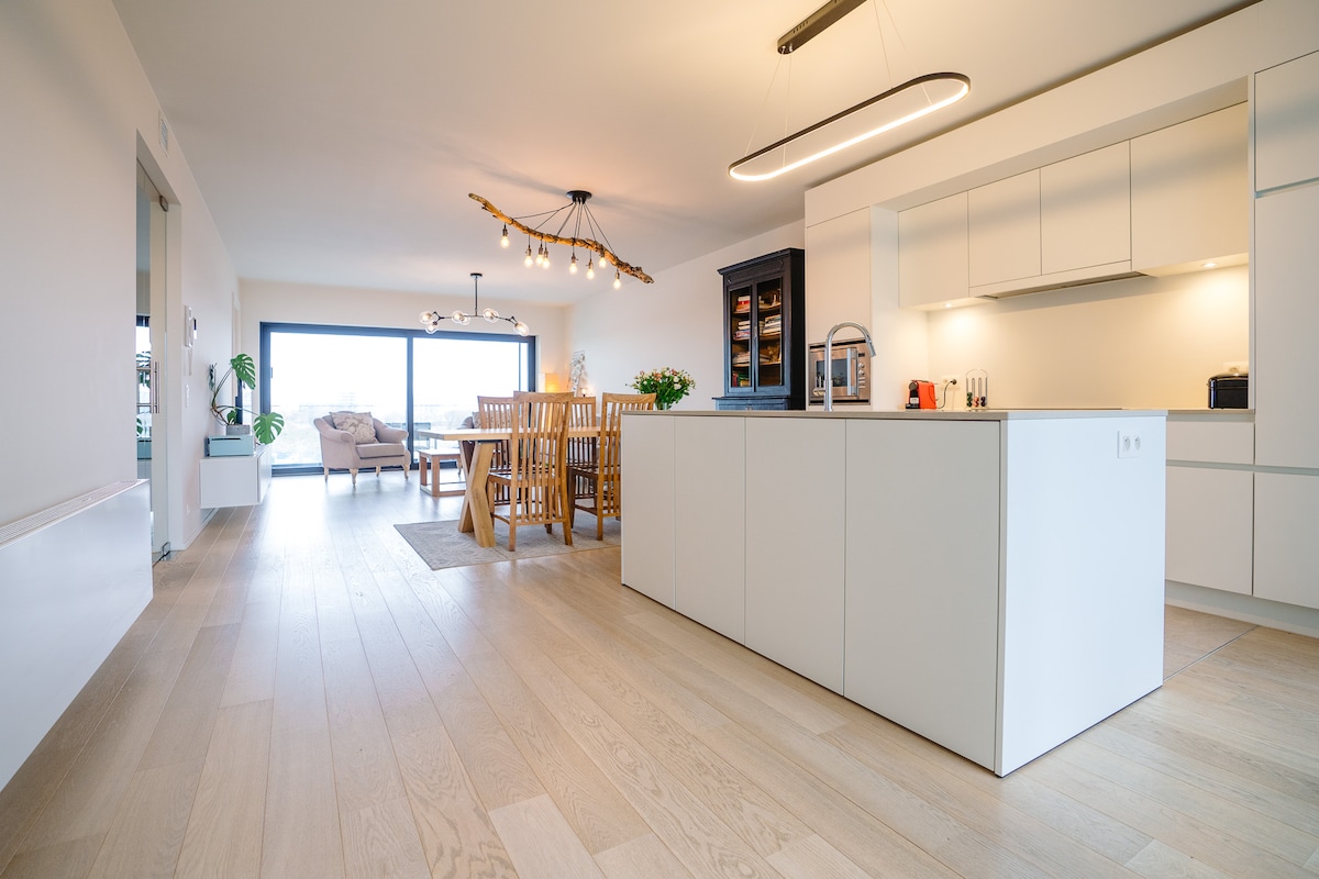 The spacious living and dining area is illuminated by natural light from large windows. A modern kitchen with an island and sleek cabinetry is visible, complemented by a dining table surrounded by wooden chairs. A cozy sitting area is also present, featuring comfortable furnishings.