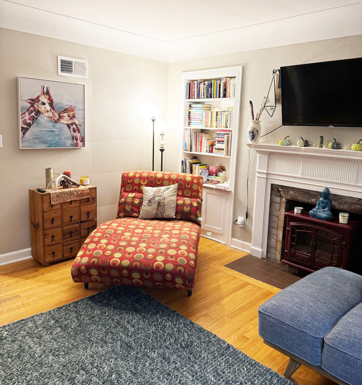 A cozy living area features a red and beige polka dot chaise lounge, accompanied by a blue upholstered chair. A decorative painting of giraffes hangs on the wall, while a bookshelf filled with books is positioned nearby. A small wooden cabinet and a fireplace add charm to the space.