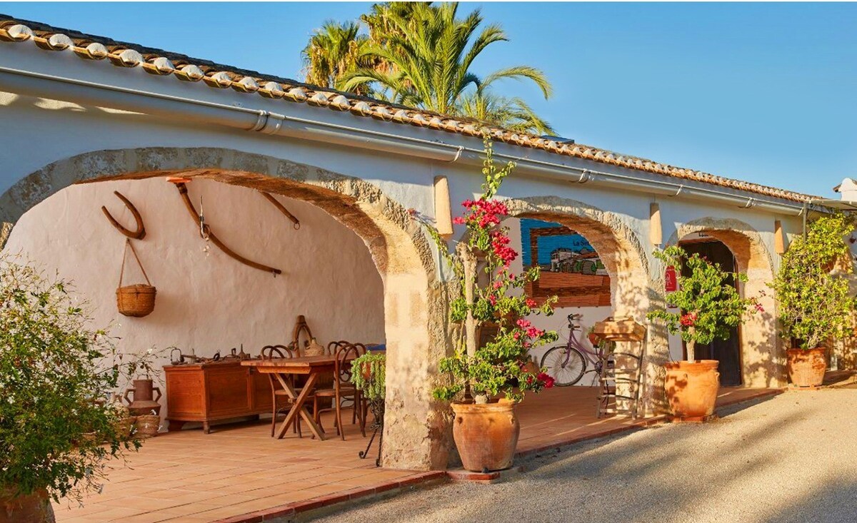 The exterior features an arched porch with terracotta flooring and wooden furniture. Potted plants and vibrant bougainvillea enhance the rustic charm. Traditional tools hang on the wall, and a bicycle leans against the side, inviting leisurely exploration of the surrounding area.