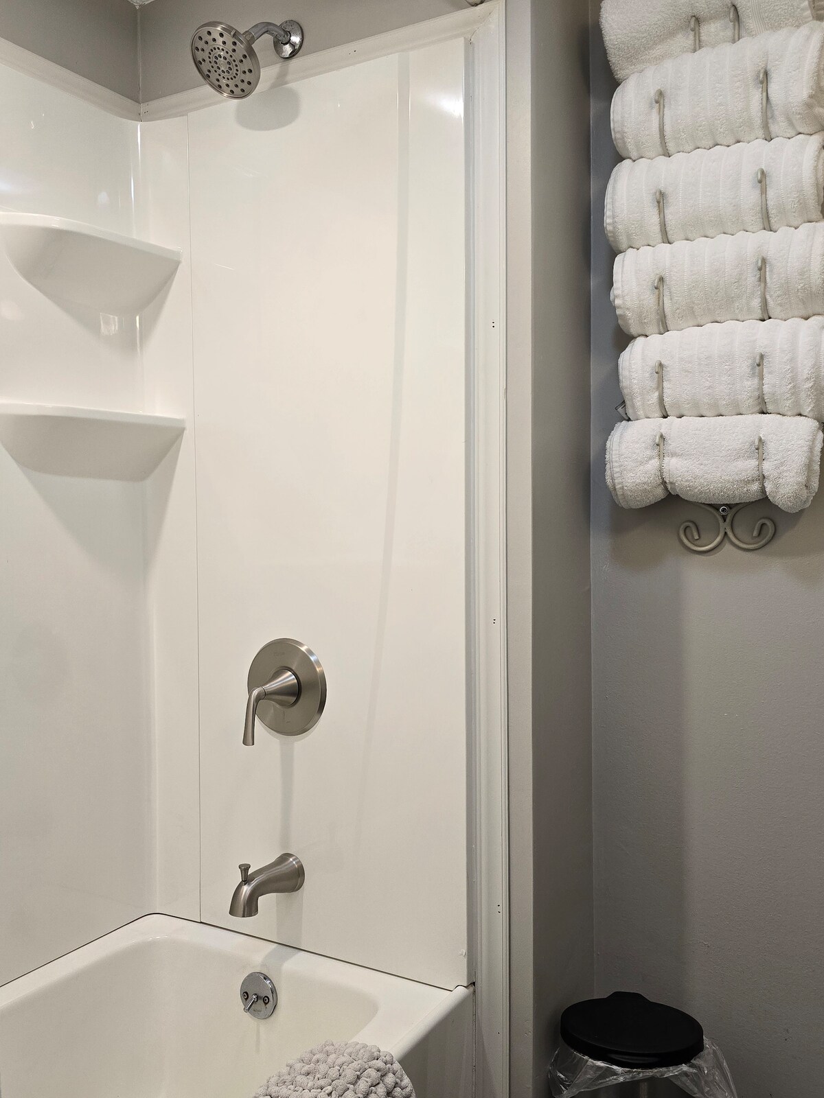 A clean and modern bathroom features a white tub and shower combination. An overhead showerhead is present, along with a polished faucet. Soft, neatly arranged towels hang on a rack nearby, while a wastebasket is visible on the floor.