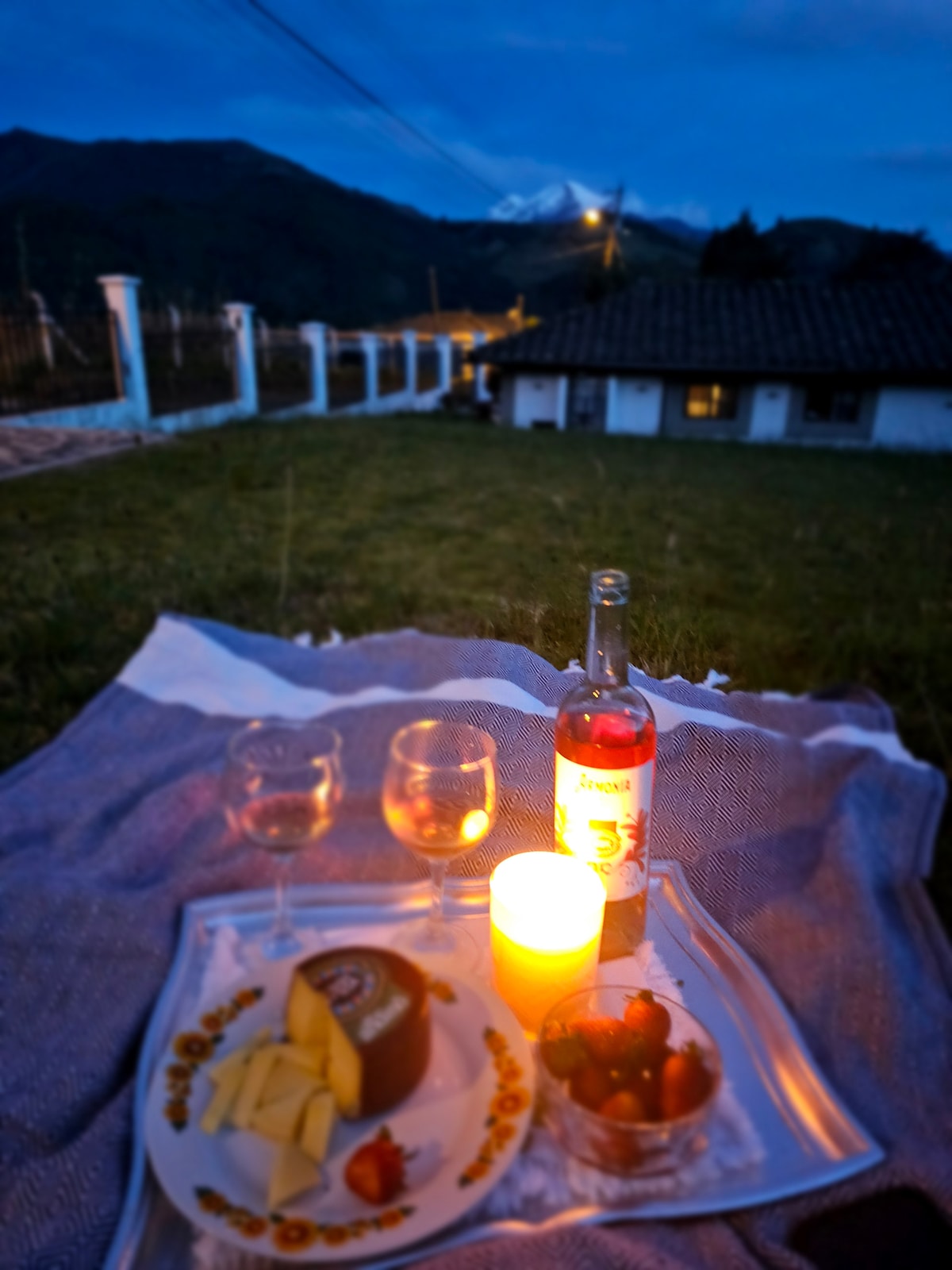 A cozy outdoor scene is set with a blanket and a tray featuring glasses, a bottle of wine, cheese, and strawberries. A glowing candle adds warm light, while the mountains are visible in the background against the evening sky.