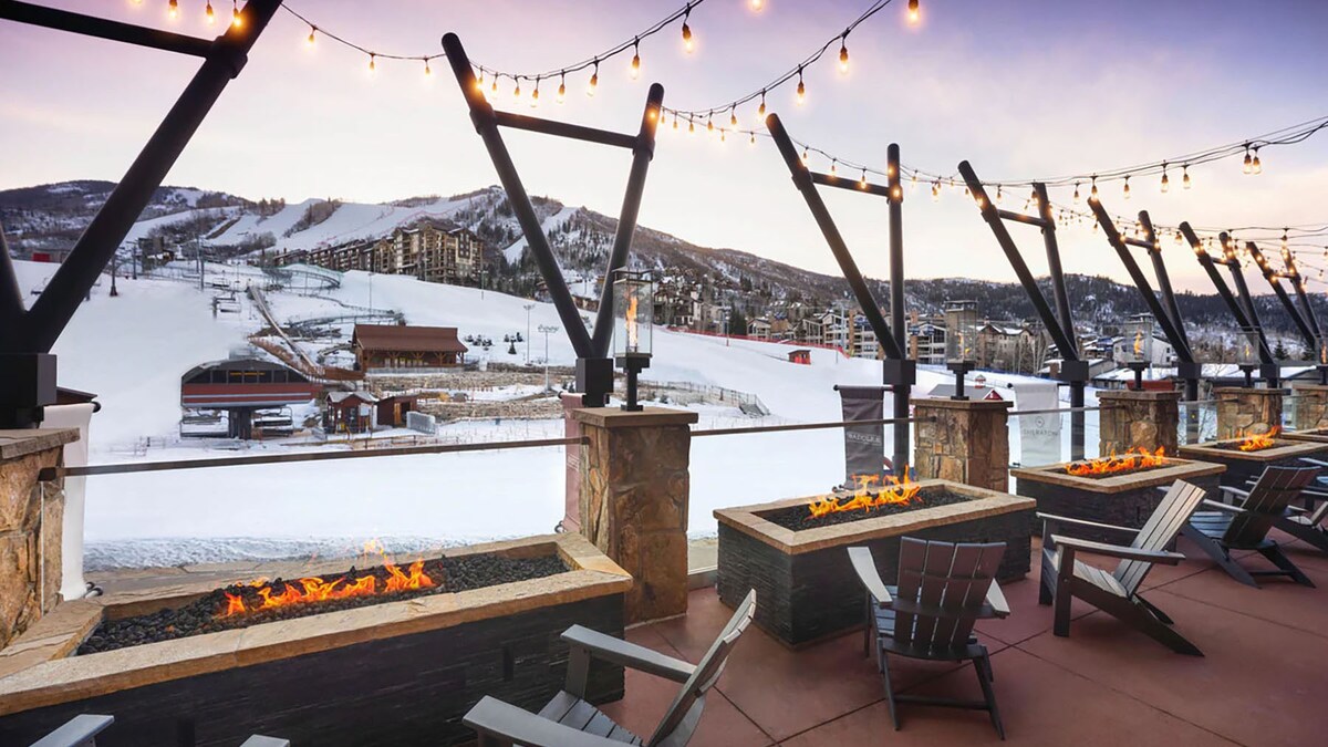An expansive outdoor terrace features multiple fire pits surrounded by lounge chairs, providing a warm gathering space. Strung lights add ambiance against the backdrop of snow-covered slopes and the distant ski facilities.