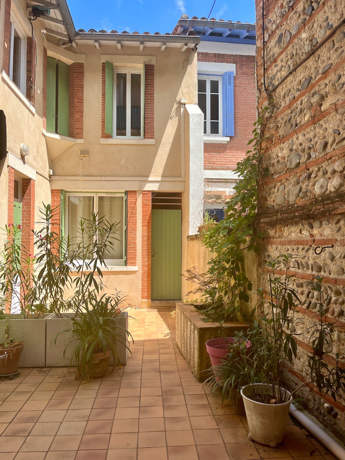 A tranquil courtyard is presented, featuring terracotta tiles and greenery. Surrounding walls display a mix of brick and stone textures. Soft natural light illuminates the space, highlighting the green door and windows adorned with shutters. Potted plants are arranged along the pathway.