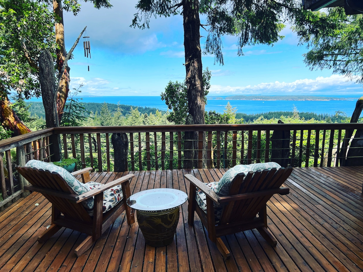 A wooden deck overlooks the expansive water view of the Georgia Strait. Two comfortable Adirondack chairs are positioned to allow for relaxation while enjoying the scenery. Trees frame the scene, adding a sense of seclusion and tranquility.