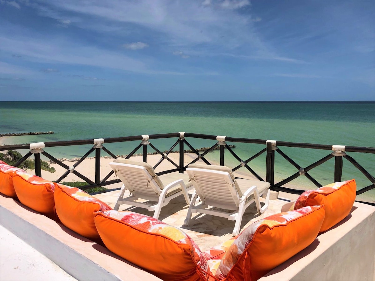 A rooftop terrace features two lounge chairs facing the Gulf, offering unobstructed views of the ocean and sandy beach. Colorful cushions line the railing, providing a comfortable seating area for relaxation. The blue sky and calm waters create a peaceful backdrop.