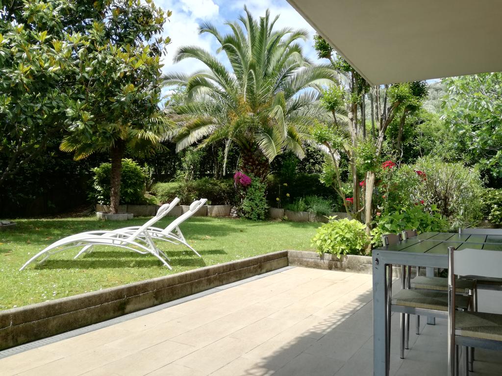 A private garden is depicted, featuring well-kept grassy areas and tropical vegetation. Two lounge chairs are positioned on a stone patio, with a table and chairs nearby. Lush palms and blooming flowers add to the tranquil setting, inviting relaxation and outdoor enjoyment.