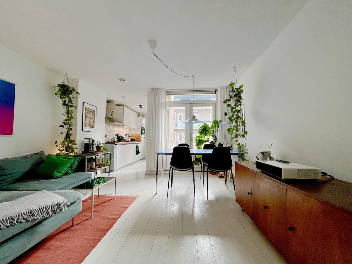 A cozy living space features a green sofa with decorative cushions, accompanied by a small table set for two. Natural light floods in through a large window, illuminating indoor plants that add a touch of greenery. A television sits atop a wooden cabinet, enhancing the room's functionality.