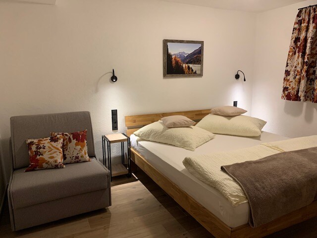 A cozy sleeping area features a spacious bed with plush pillows and a warm throw blanket. Next to it, a small nightstand holds a reading lamp. A comfortable chair with decorative cushions is positioned nearby, and a nature-themed artwork adorns the wall.