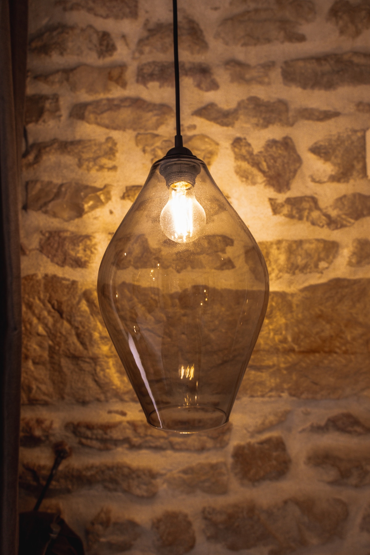 A pendant light fixture is suspended from a ceiling, featuring a clear glass shade that gently curves. The exposed bulb emits a warm glow, illuminating the textured stone wall behind, enhancing the cozy ambiance of the space.
