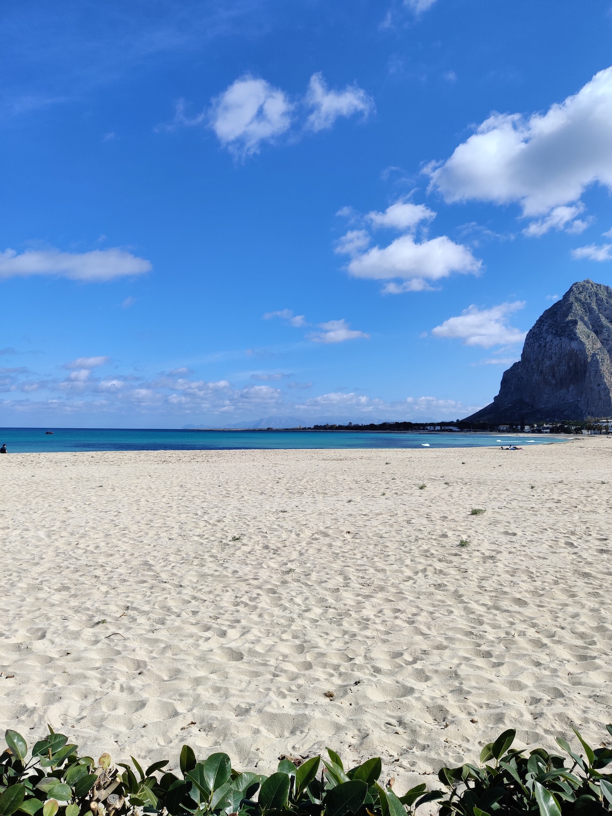 A wide sandy beach extends towards a clear blue sea under a partly cloudy sky. In the distance, a rocky cliff rises prominently, creating a striking contrast against the water. Sparse vegetation is visible in the foreground, framing the serene landscape.