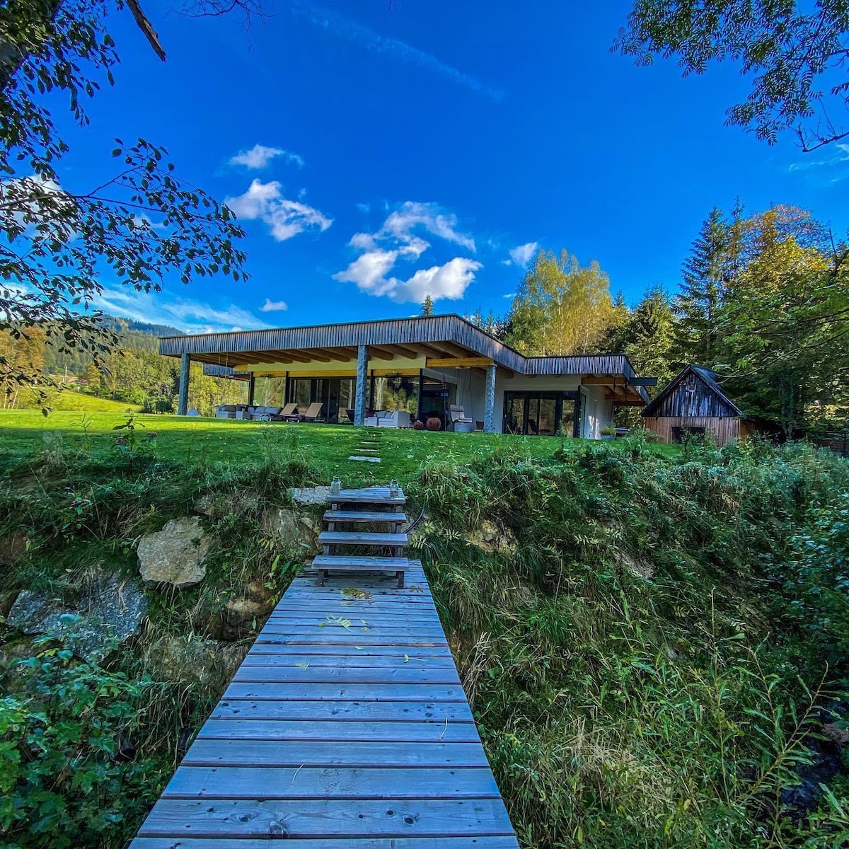 A modern retreat is showcased with a flat roof and large panoramic windows nestled among trees. A wooden walkway leads from a natural area to the spacious outdoor patio, encouraging relaxation and connection to the surrounding landscape under a clear blue sky.