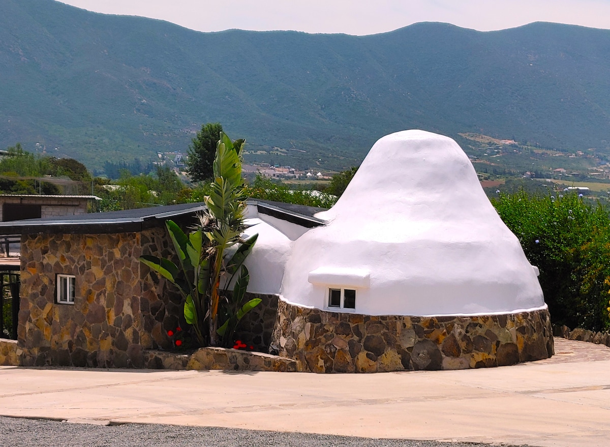 The unique Eco-Dome Home showcases a distinctive rounded structure with a smooth, white exterior. A stone wall surrounds the home, complemented by green foliage and tropical plants. The backdrop features rolling mountains, contributing to the serene landscape of Valle de Guadalupe.