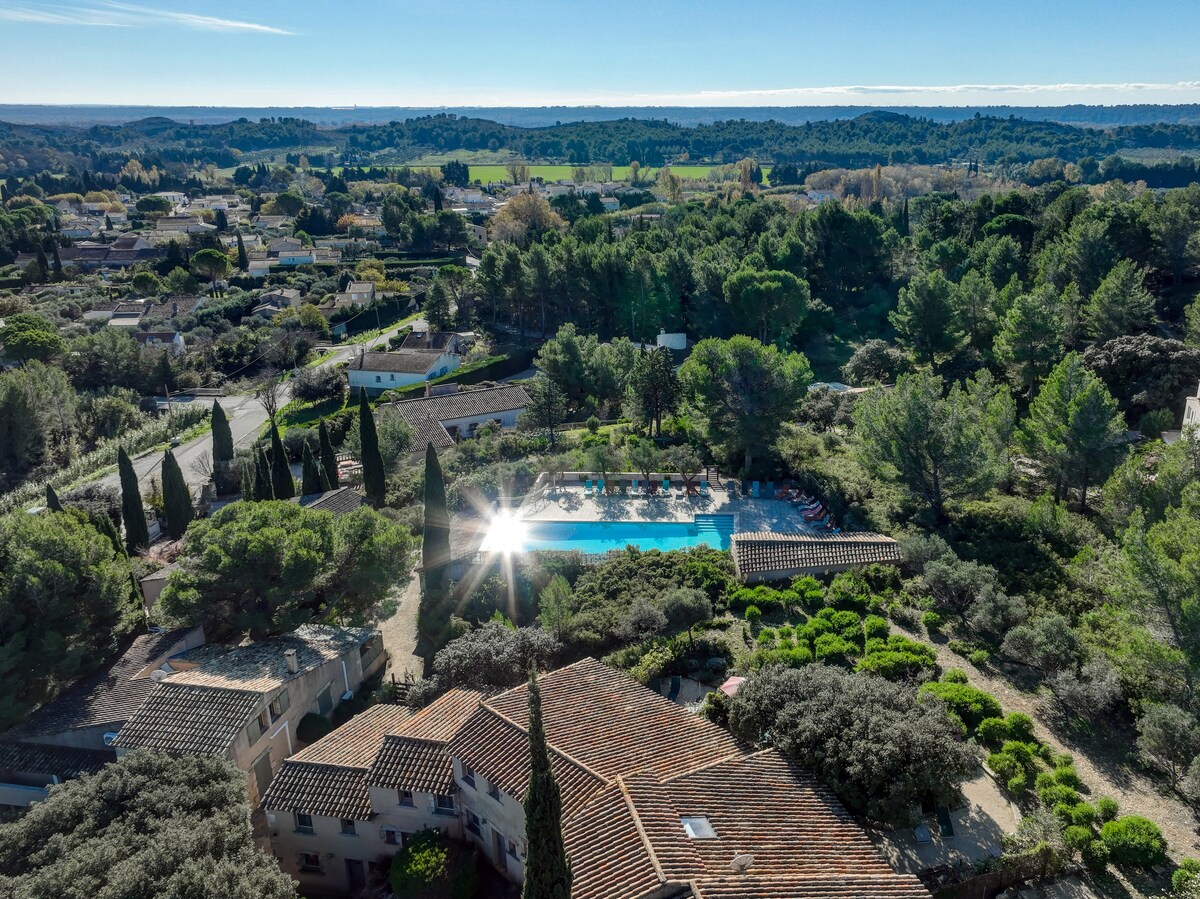 A panoramic view showcases a serene landscape of lush greenery and a shared swimming pool. The pool is surrounded by trees and landscaped gardens, reflecting sunlight and creating a peaceful ambiance. The distant hills add to the tranquil setting of the vacation residence.