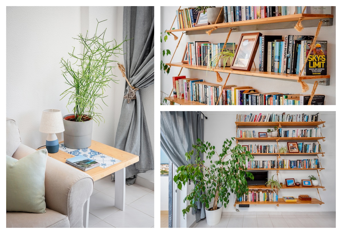 Stylish and modern living space features a neutral colour palette complemented by green plants. A wooden coffee table with a potted plant and lamp is positioned next to a comfortable sofa. A wall-mounted bookshelf holds a variety of books, enhancing the room's inviting atmosphere.