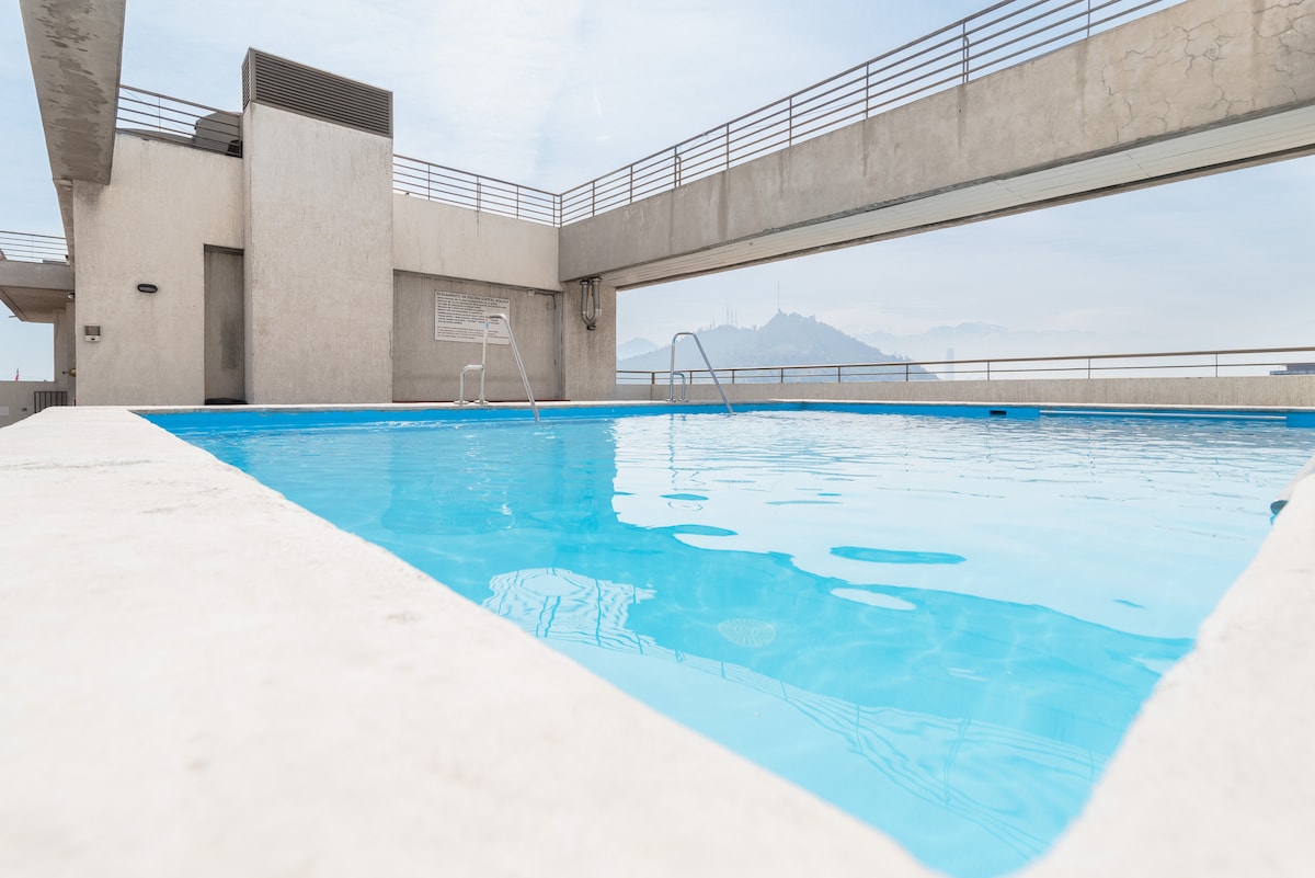 A rooftop pool is visible, featuring clear blue water and a simple ladder for access. Surrounding areas consist of concrete walls and railings, with distant views of mountains in the background. The overall setting is bright and open, offering ample space for enjoyment.
