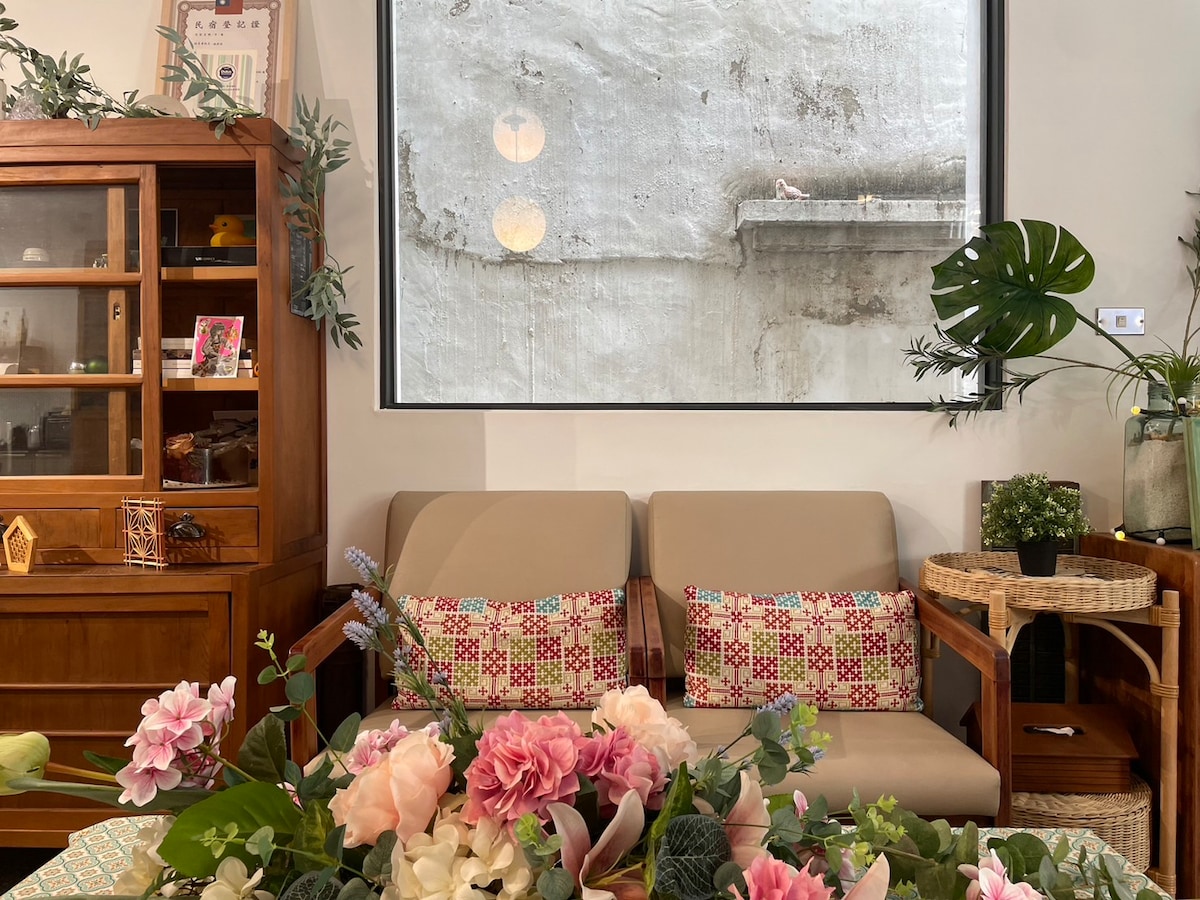 A seating area features two cream-colored sofas with decorative cushions, positioned in front of a large window showcasing a textured concrete wall. A table adorned with an array of colorful flowers is visible, complemented by wooden furniture and greenery placed nearby.
