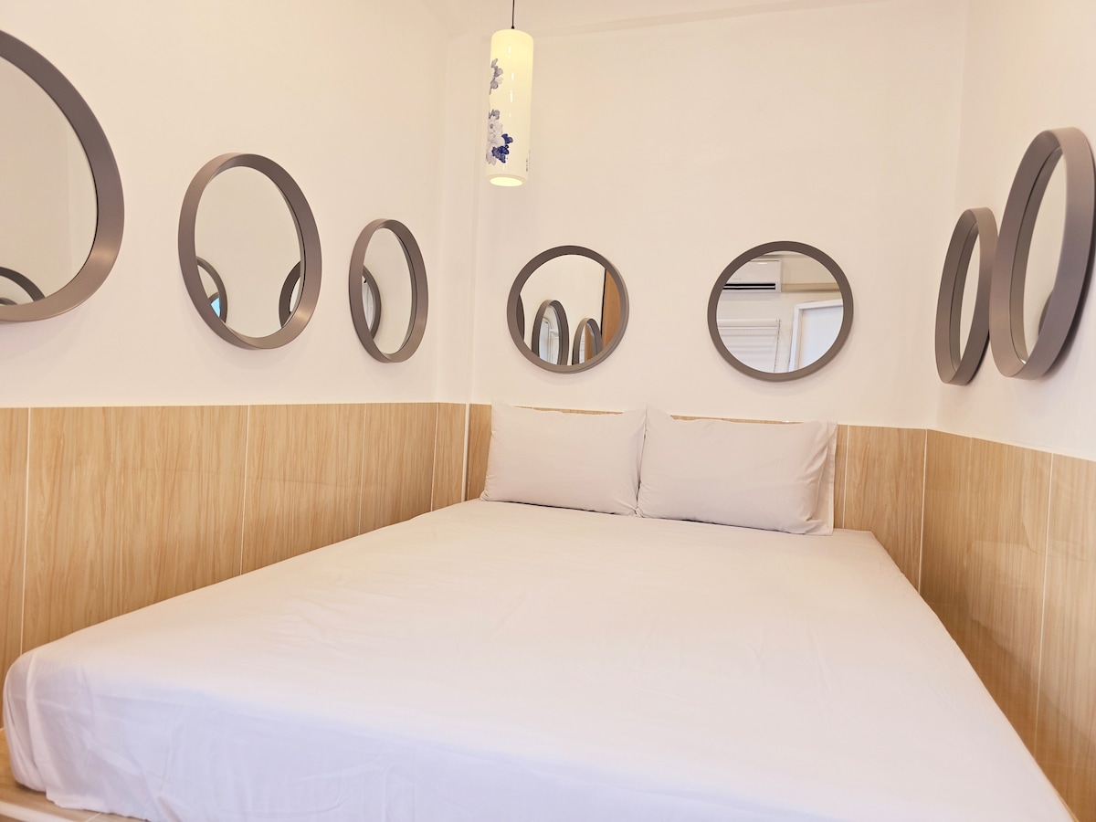 A minimalist sleeping area features a spacious bed covered with a fitted white sheet. Multiple round mirrors adorn the walls, reflecting light. The warm wood paneling complements the bright white walls and ceiling, creating a simple yet cozy atmosphere.