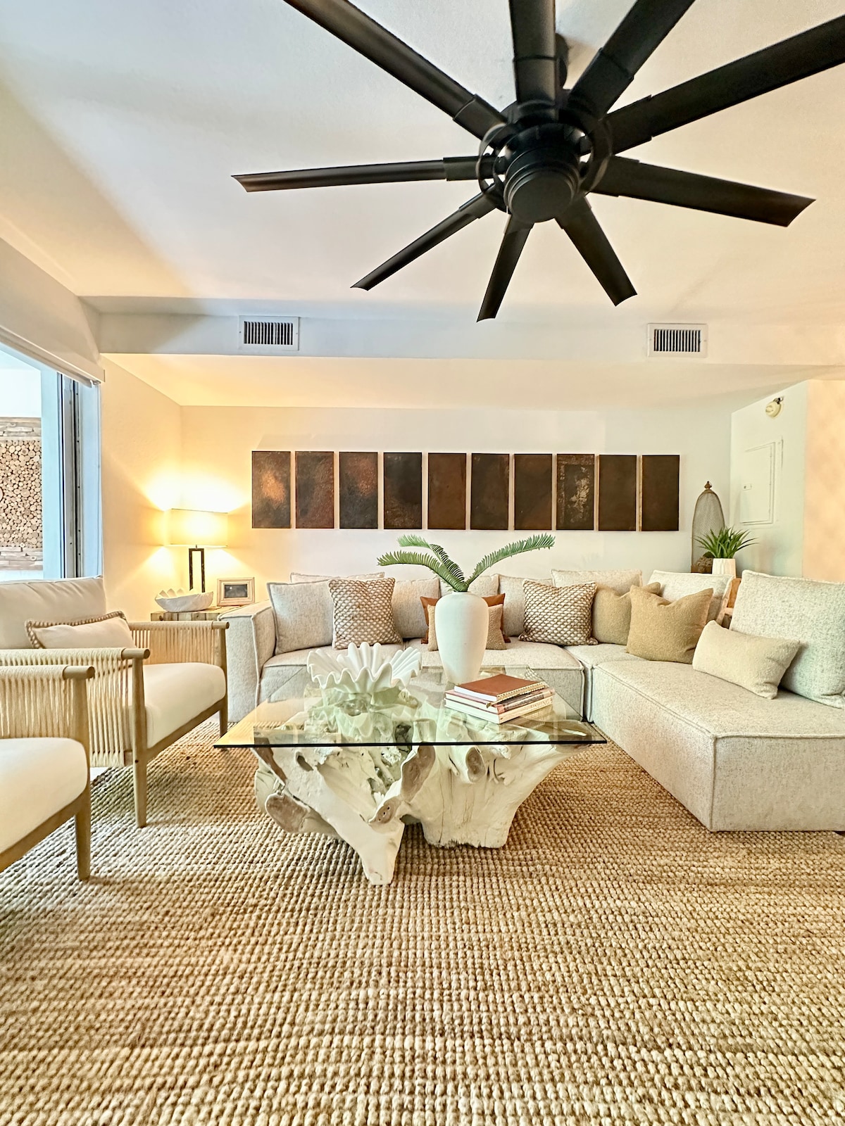 A spacious living area is highlighted by a large sectional sofa adorned with neutral-toned cushions. A coffee table made from natural wood sits at its center. Soft lighting is provided by a floor lamp, while a ceiling fan adds airflow. Minimalist art pieces adorn the wall.
