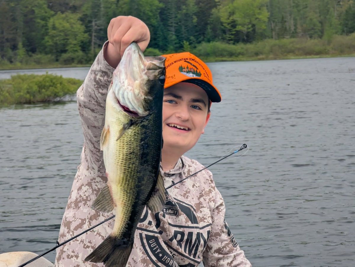 A person holds a large bass, showcasing its vibrant green and white coloring. The background features a calm pond surrounded by lush greenery, embodying the tranquil setting of Twin Ponds Cottage's fishing opportunities.