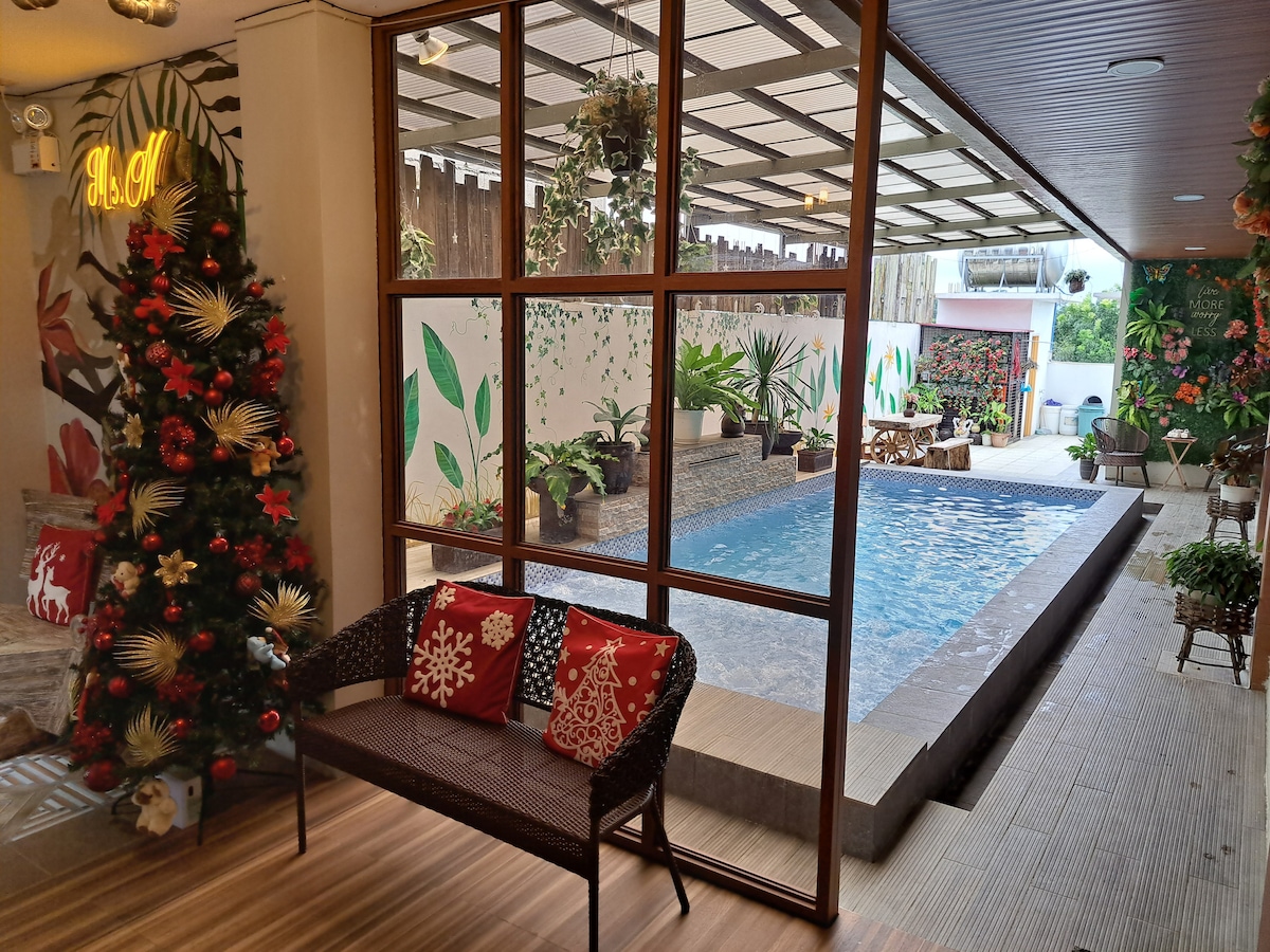 A spacious indoor infinity pool is visible through large glass windows, surrounded by lush greenery and vibrant murals. A seating area with decorative pillows is positioned nearby, creating a comfortable spot to enjoy the view. Holiday decorations add a festive touch to the space.