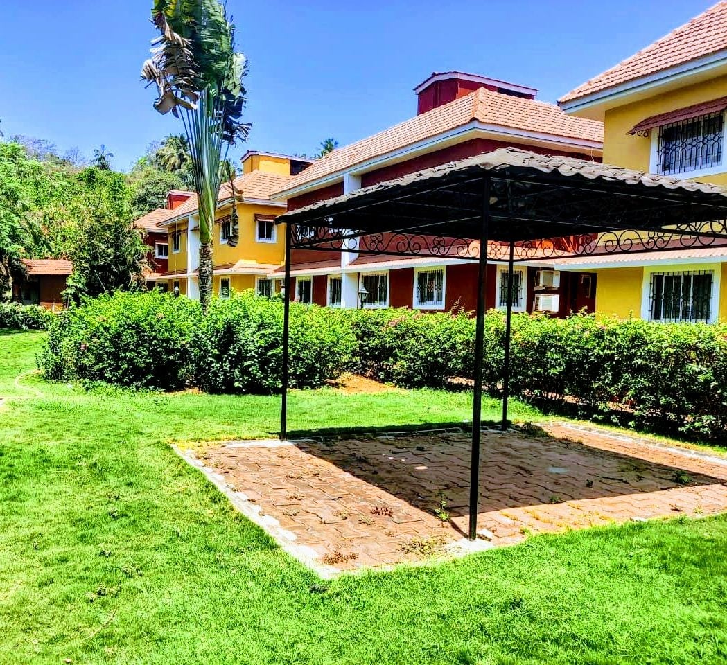 A shaded gazebo is situated on a grassy area surrounded by manicured shrubs and plants. The background features several Portuguese styled villas, characterized by vibrant colors and tiled roofs, set against a clear blue sky.