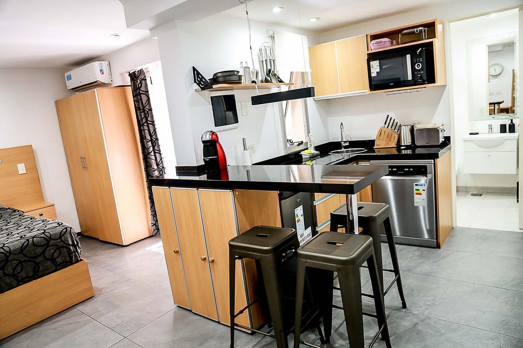 The studio features a modern kitchen with a large island, black countertops, and stainless steel appliances. Seating is provided at three high stools. A cozy sleeping area is visible in the background, with a double bed and light-colored furniture. Bright light fills the space, enhancing its contemporary design.