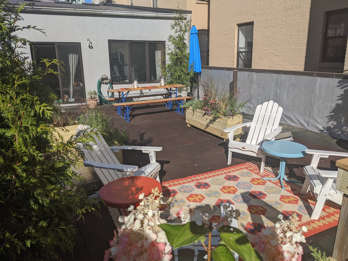 A private terrace is featured with wooden lounge chairs and small tables, surrounded by planters filled with greenery and flowers. A colorful area rug lies on the ground, while a large blue umbrella provides shade beside a long communal table.
