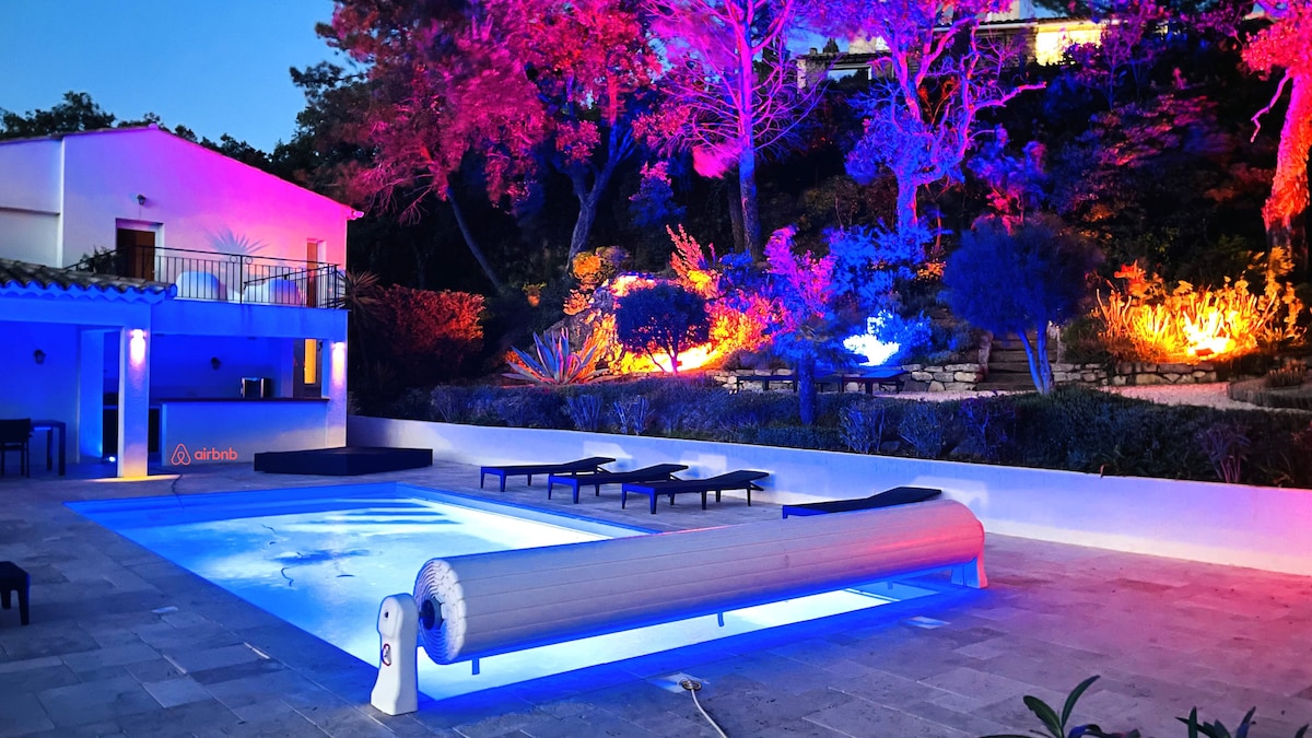 A well-lit outdoor area features a swimming pool surrounded by lounge chairs. The pool's surface reflects vibrant hues created by illuminated landscaping, while a white wall and a light-colored villa provide a serene backdrop. Subtle foliage enhances the peaceful atmosphere of the space.