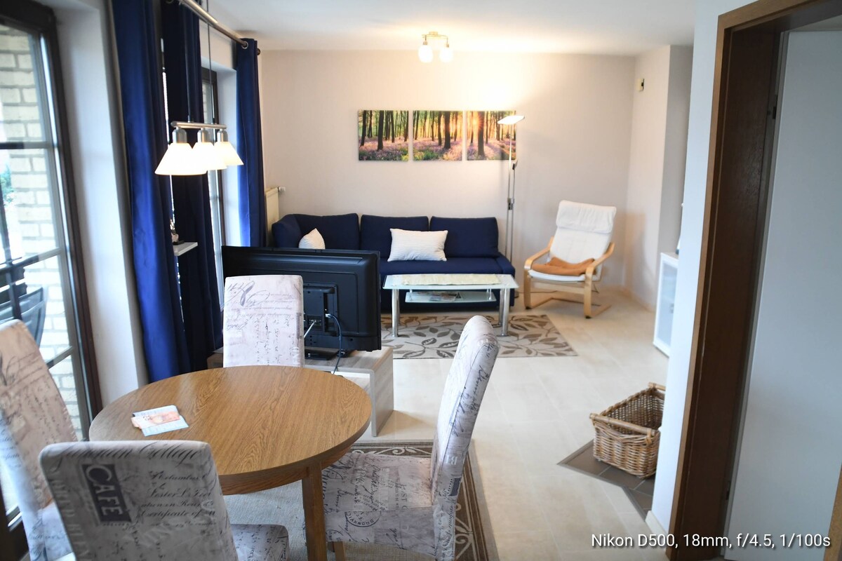 A dining area features a round wooden table surrounded by two chairs, with a cozy seating arrangement visible in the background. A television is positioned nearby, and natural light illuminates the space through large windows, adding a bright and airy feel.