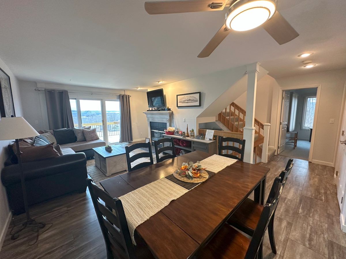 A dining area features a dark wooden table with seating for six, complemented by a centerpiece of fruit. A comfortable living area is visible, showcasing a sofa and an entertainment unit, with large windows allowing natural light and valley views.