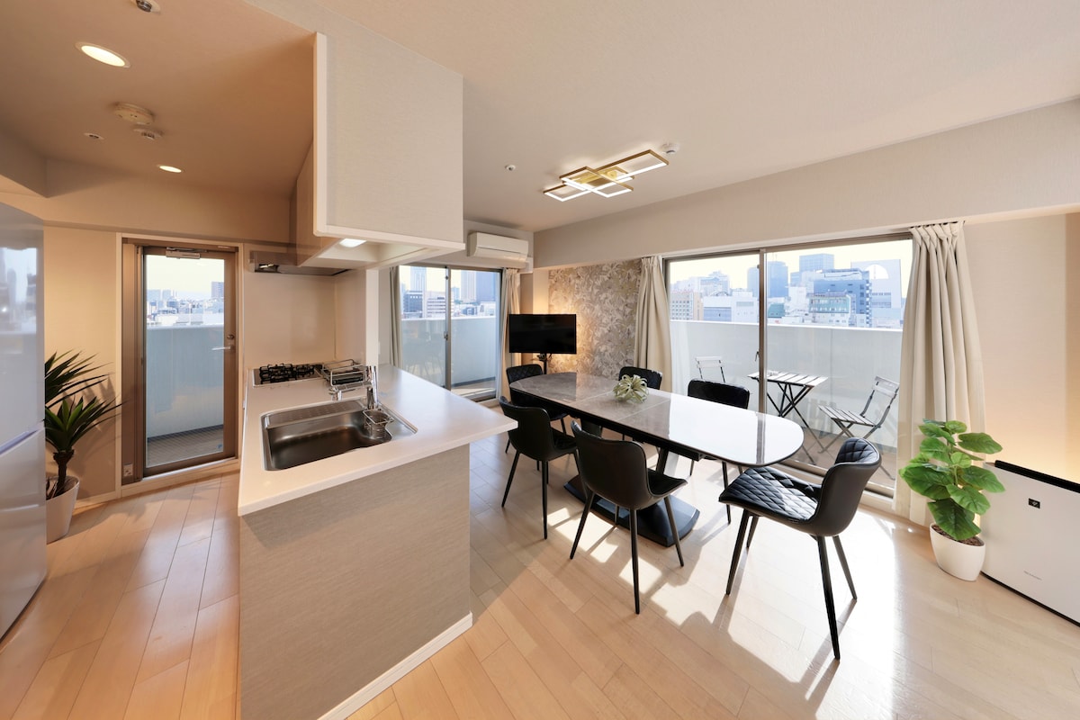 An open dining and living area features a modern kitchen with a sink and gas stovetop. A large dining table is surrounded by black chairs. Floor-to-ceiling windows offer city views, and a small balcony with outdoor seating is visible. Potted plants add a touch of greenery.