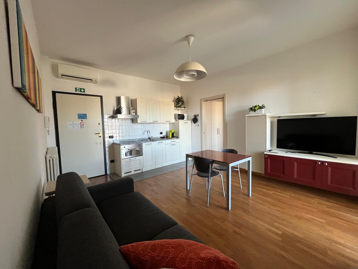 A light-filled living area features a modern kitchenette with white cabinetry and essential appliances, including a refrigerator and stove. A dining table with four chairs is positioned nearby, and a comfortable sofa is placed opposite a television unit with storage. Air conditioning is visible on the wall.