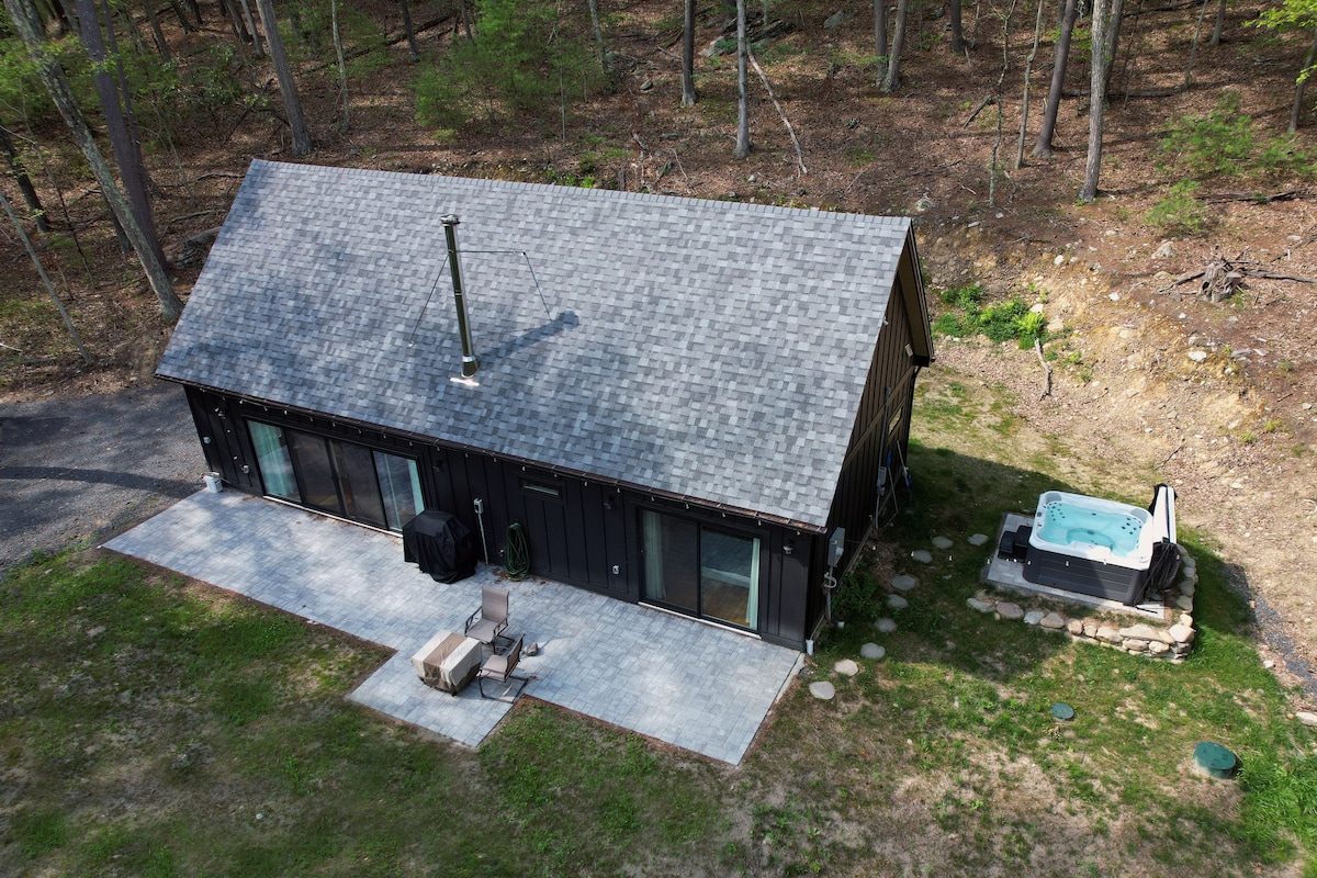 The modern cabin is set within a forested area, featuring a sloped roof and large windows. A private patio is furnished with a seating area and a hot tub is positioned nearby, surrounded by stone. The natural landscape enhances the serene setting.
