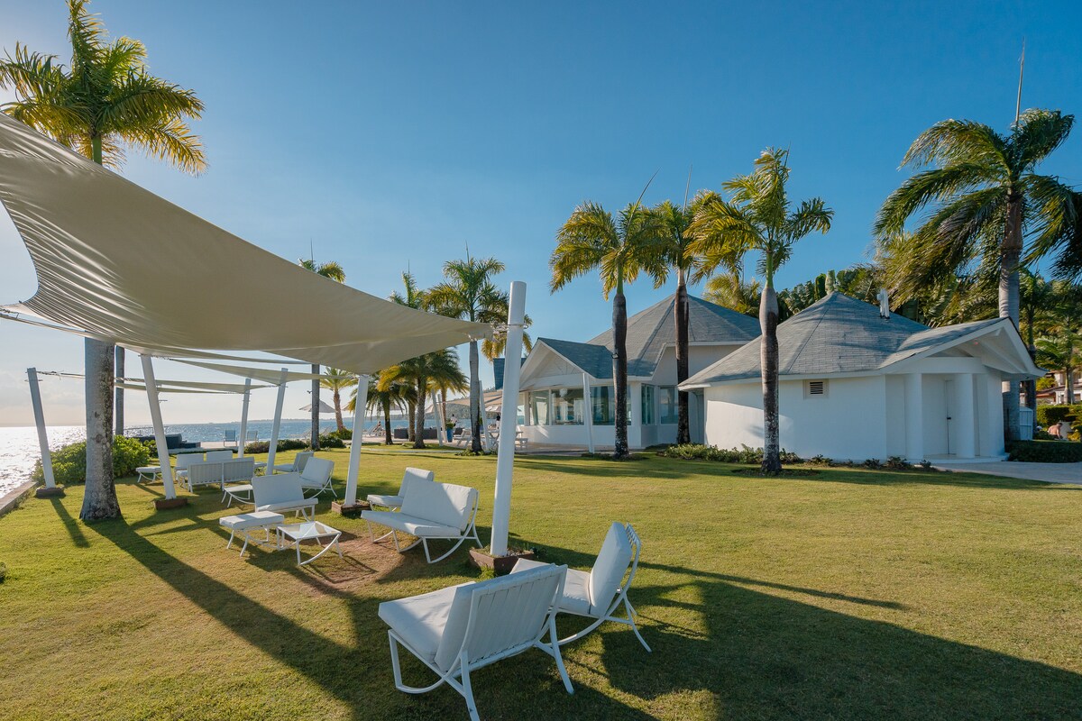 An outdoor area features several lounge chairs positioned on a well-manicured lawn. A large shade sail provides coverage against the sun, while palm trees gently sway in the breeze. The serene sea is visible in the background, enhancing the tranquil atmosphere.