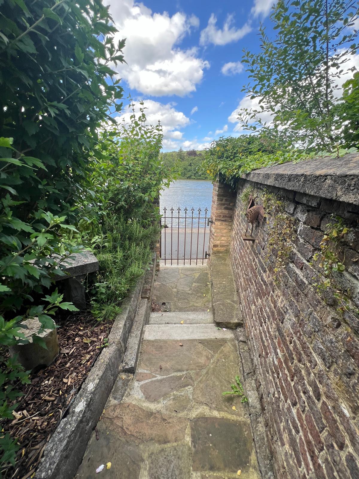 A stone pathway leads through lush greenery and a wrought iron gate, opening to a serene view of the water. The rustic brick wall frames the scene, with the sky visible above, filled with fluffy clouds.