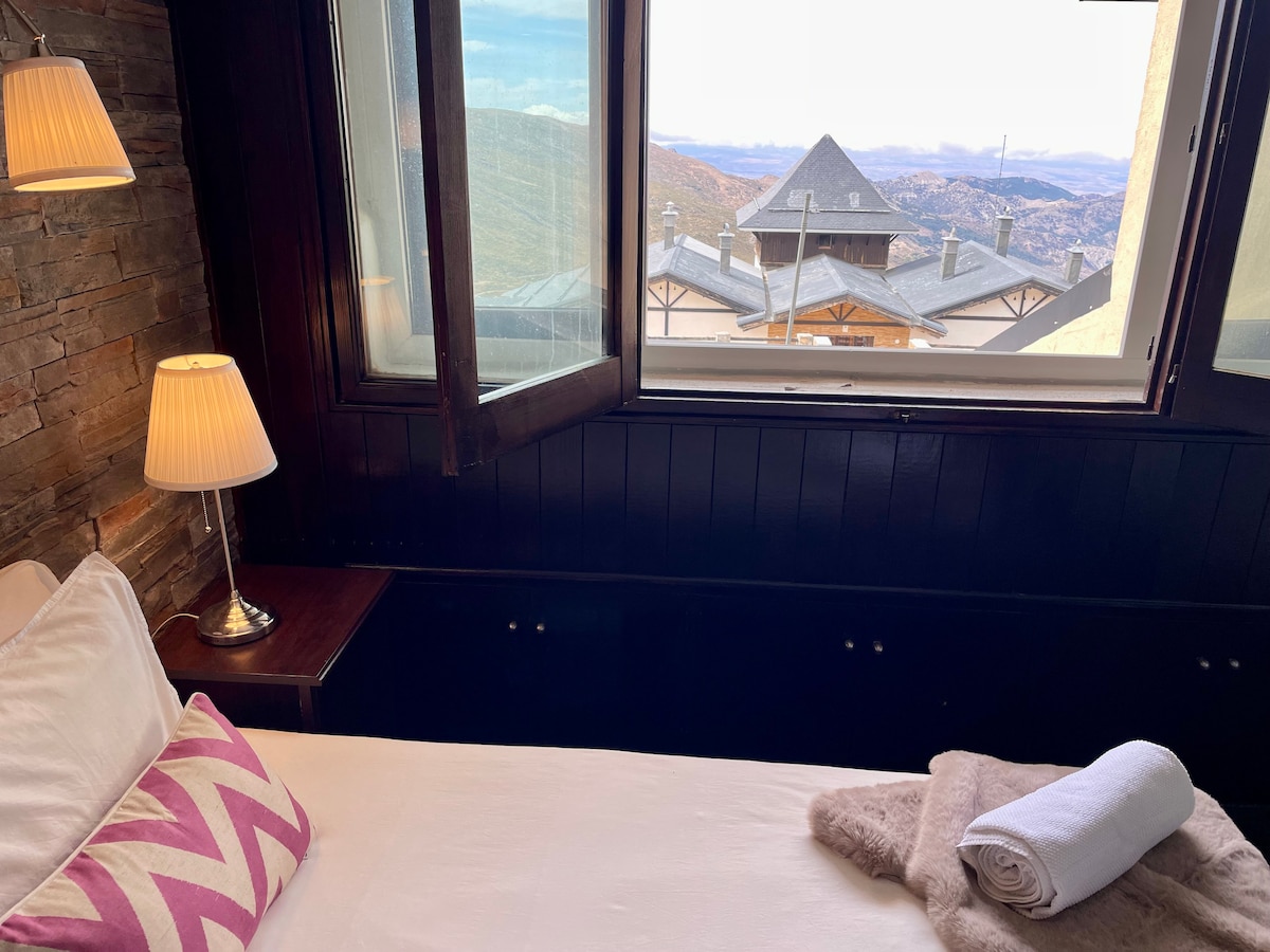 A bright bedroom features a spacious bed adorned with soft linens and decorative pillows. Natural light pours in through large windows, offering views of the mountainous landscape. A bedside lamp stands next to the bed, complementing the warm tones of the wooden and stone details.