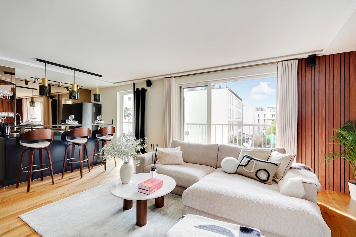 A modern living space features a spacious sectional sofa adorned with decorative cushions. A round coffee table sits centrally, complemented by a neutral area rug. The open kitchen area is visible in the background, showcasing stylish bar stools and large windows that invite natural light.