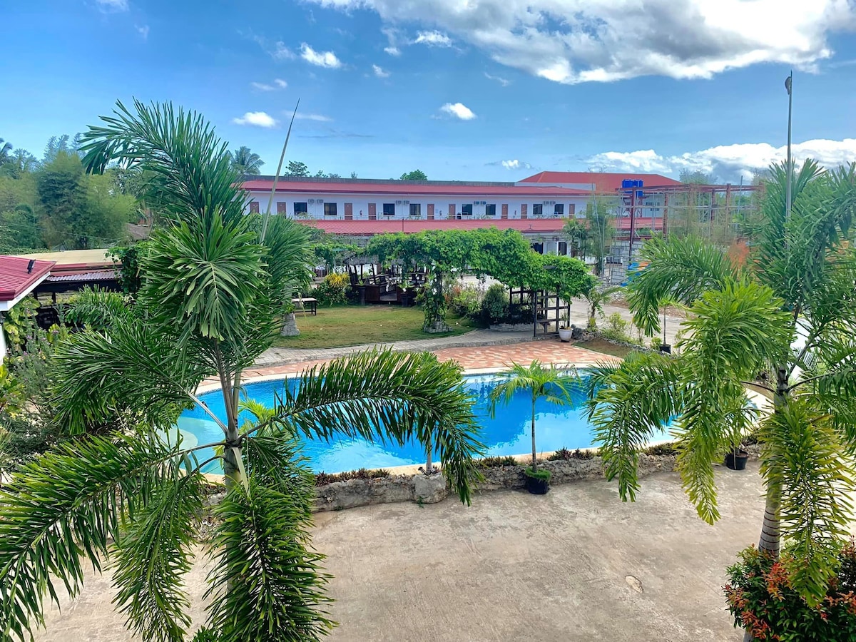 A view of the outdoor area features a swimming pool surrounded by lush greenery and palm trees. The backdrop includes a two-story building with multiple windows and a well-maintained garden area, creating an inviting atmosphere for relaxation.