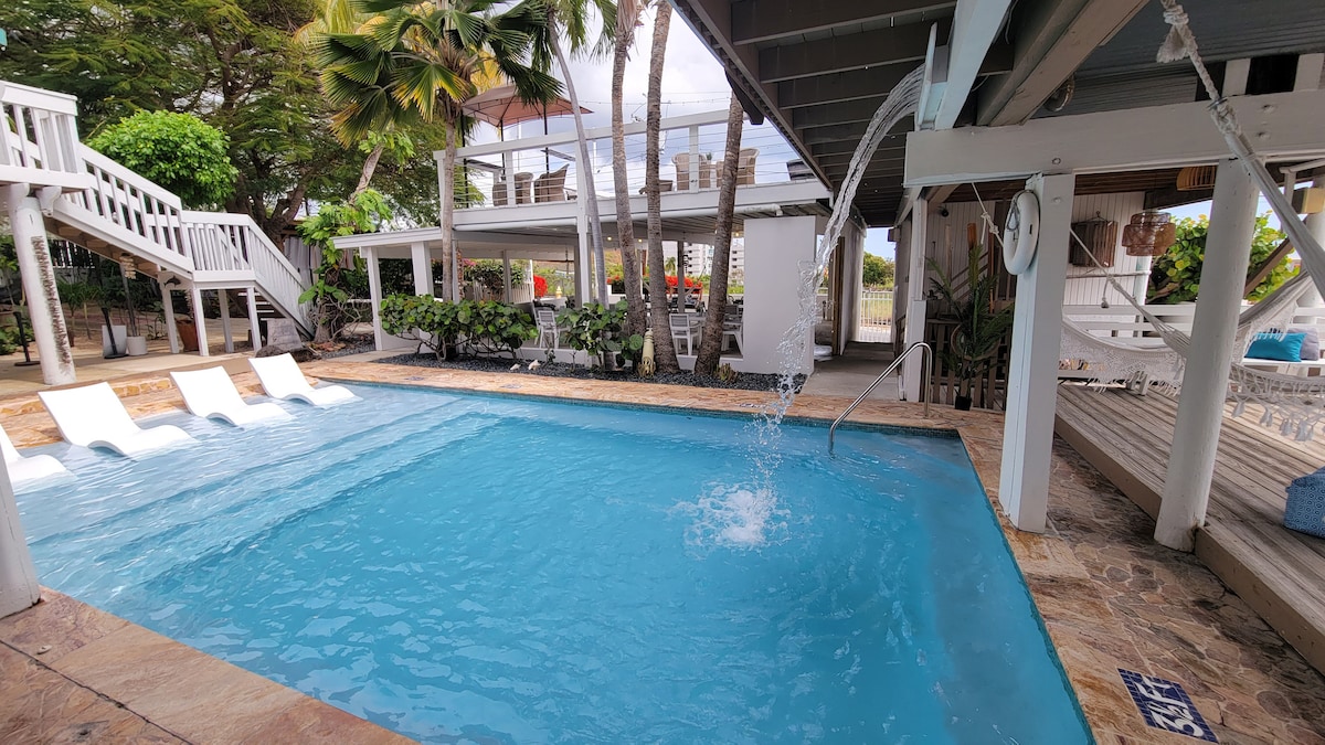 A heated swimming pool is surrounded by a stone deck, featuring four white lounge chairs. Water is seen gently splashing from a spout into the pool. Lush greenery and patio areas with varied seating options are visible in the background.