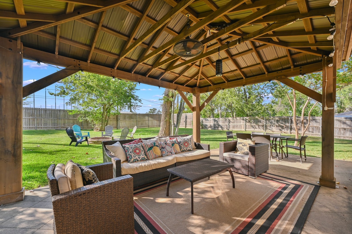 A shaded outdoor patio is featured under a wooden structure with a high roof. Comfortable seating is arranged around a central coffee table, accompanied by a patterned area rug. A dining table is positioned in the background, with lush green grass extending beyond the space.