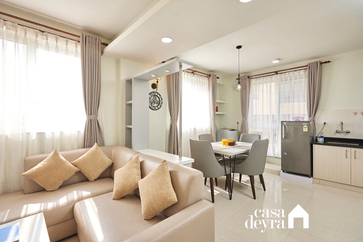 The living and dining area features a light-colored sofa adorned with decorative cushions, a dining table for four, and large windows allowing natural light. The kitchenette, equipped with modern appliances, is positioned nearby, enhancing the space's functionality.