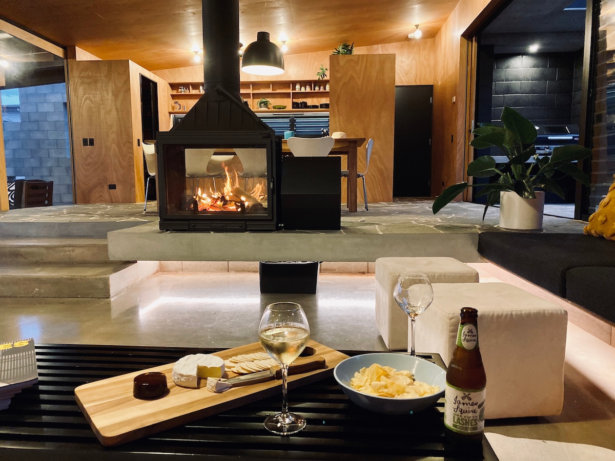 A modern living space is presented with a central fireplace, creating a focal point for relaxation. Nearby, a wooden platter holds cheese and crackers, accompanied by drinks. The open-plan area features seating, a dining table, and a functional kitchen in the background, with warm lighting enhancing the ambiance.