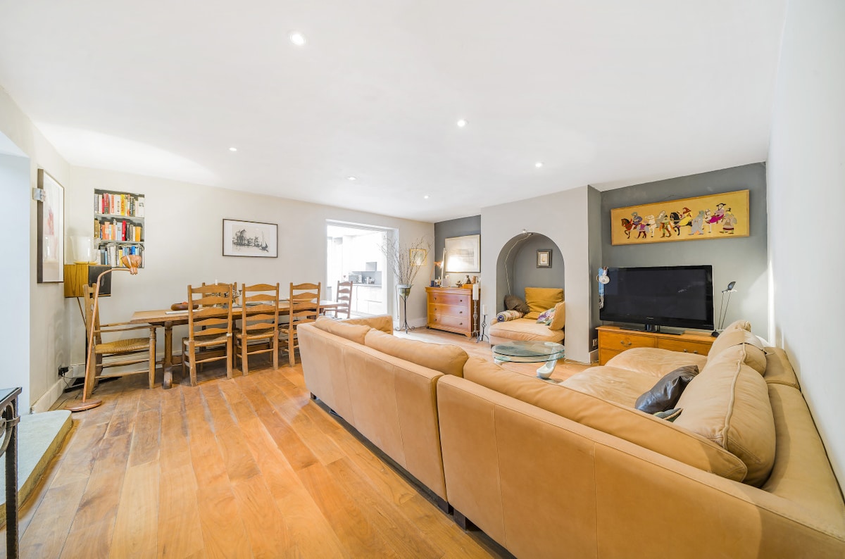 A spacious living area features a large, light-colored sofa and a wooden dining table seating six. Shelves with books are visible on one wall. A television is positioned against another wall, with soft lighting enhancing the inviting atmosphere.
