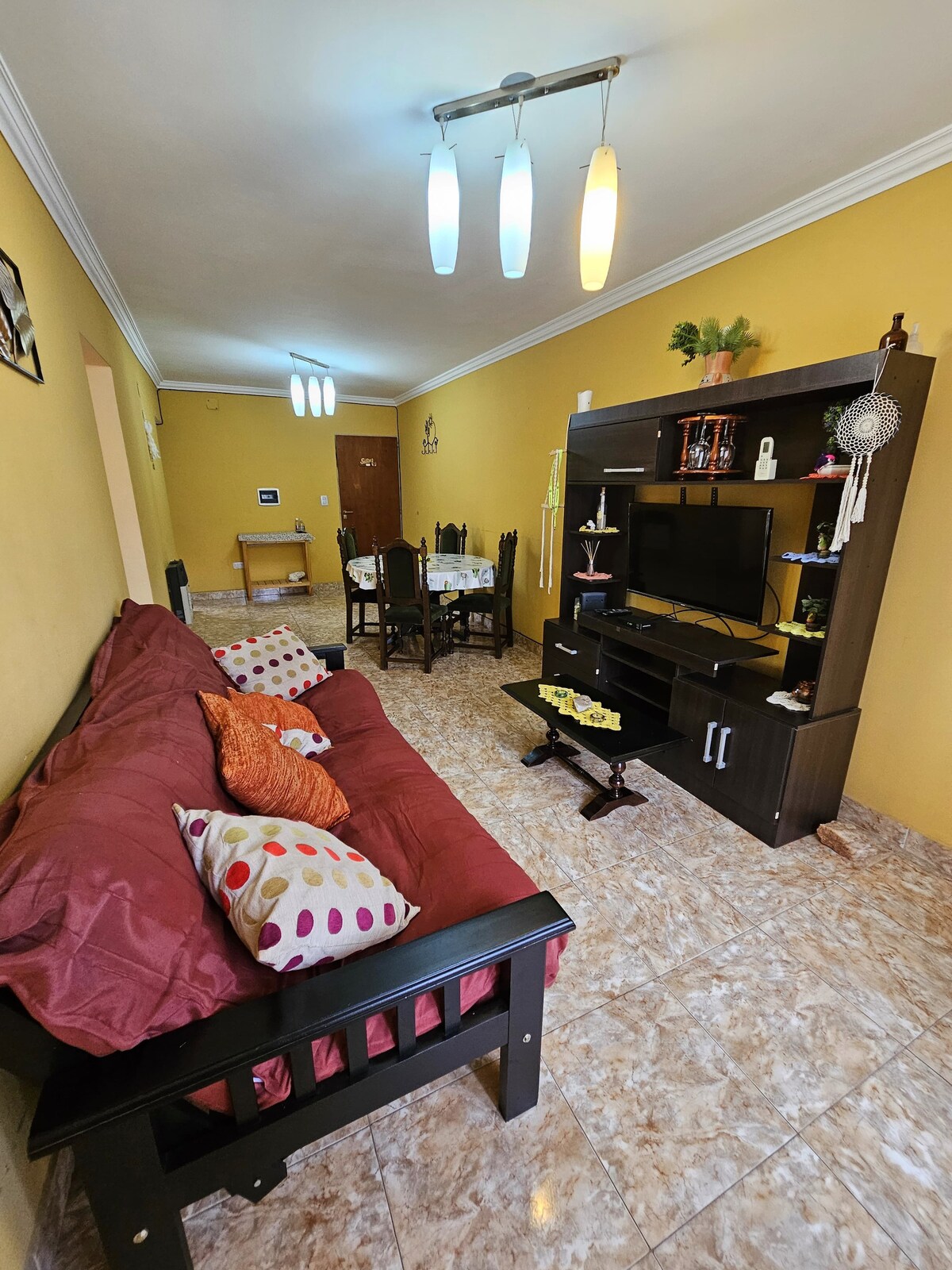 A living area is displayed with a burgundy sofa adorned with colorful cushions. A dark wood entertainment unit holds a television, surrounded by decorative items. Soft lighting fixtures hang from the ceiling, and a small dining table is visible in the background against a warm yellow wall.