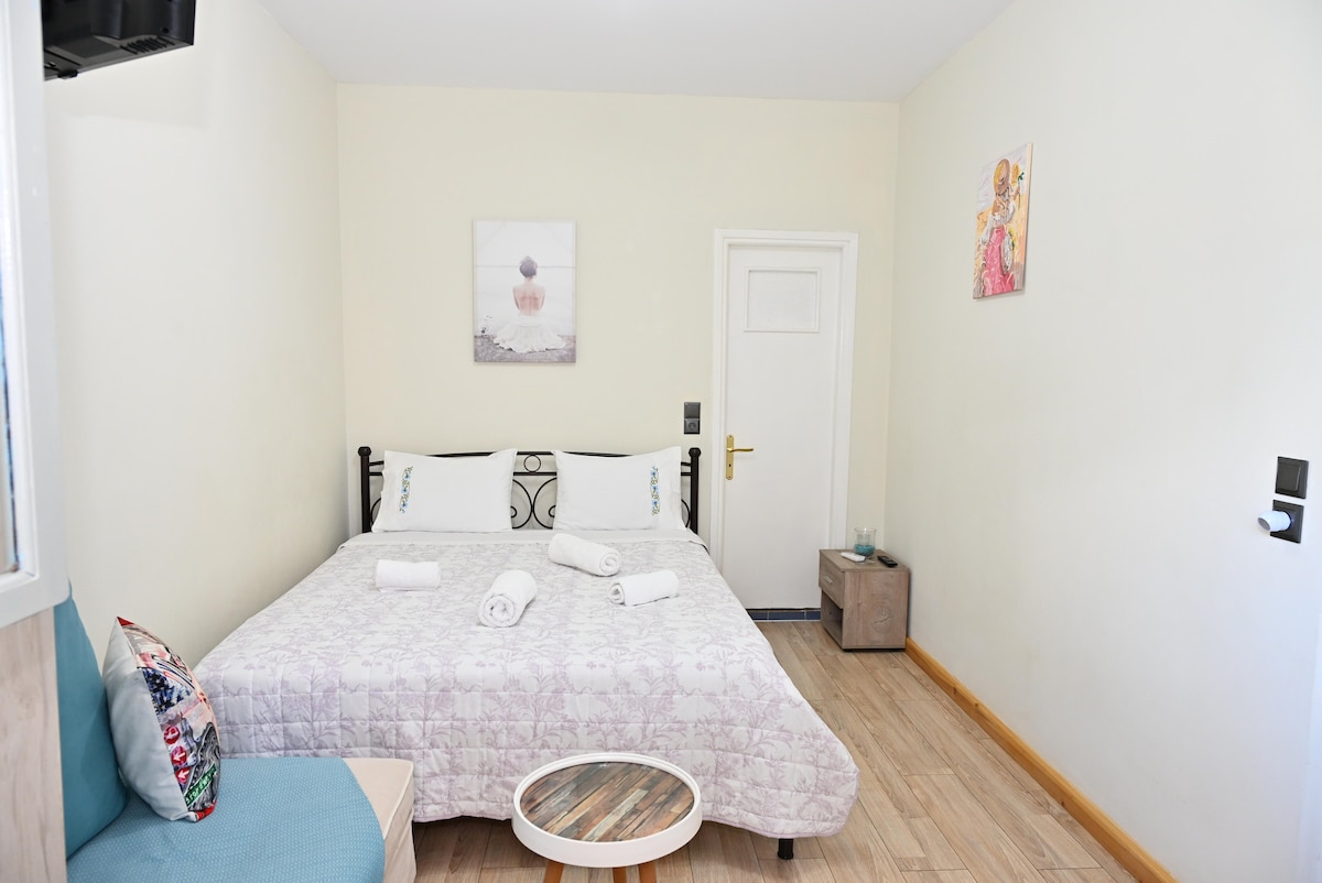 A spacious bedroom features a large bed with light bedding and white towels neatly arranged. A comfortable seating area is provided by a blue couch, while decorative wall art adds subtle charm. Natural light brightens the room through a nearby window.