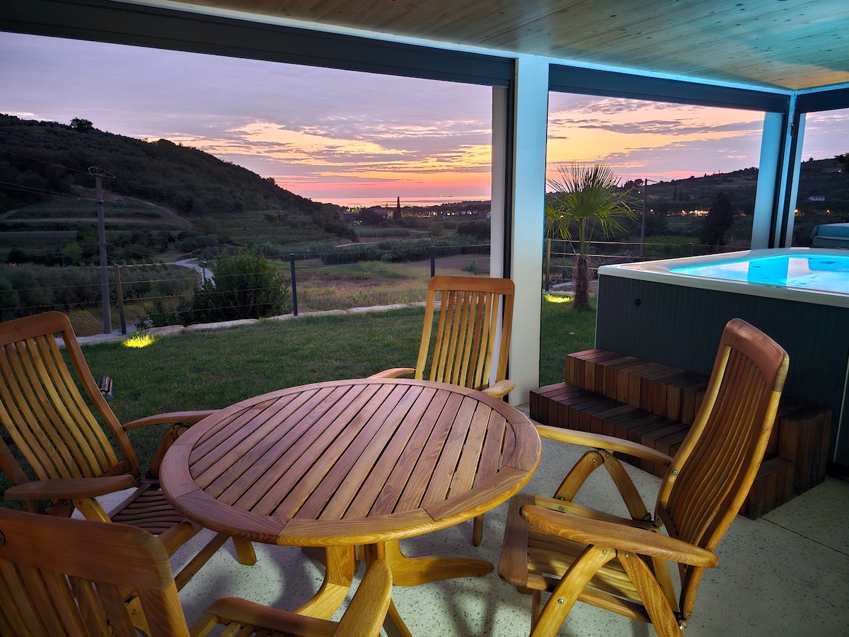 A wooden outdoor dining set, featuring a round table and four chairs, is positioned on a spacious terrace. The view offers a colorful sunset over rolling hills, enhancing the serene setting. A Jacuzzi is nearby, inviting relaxation while soaking in the scenic beauty.