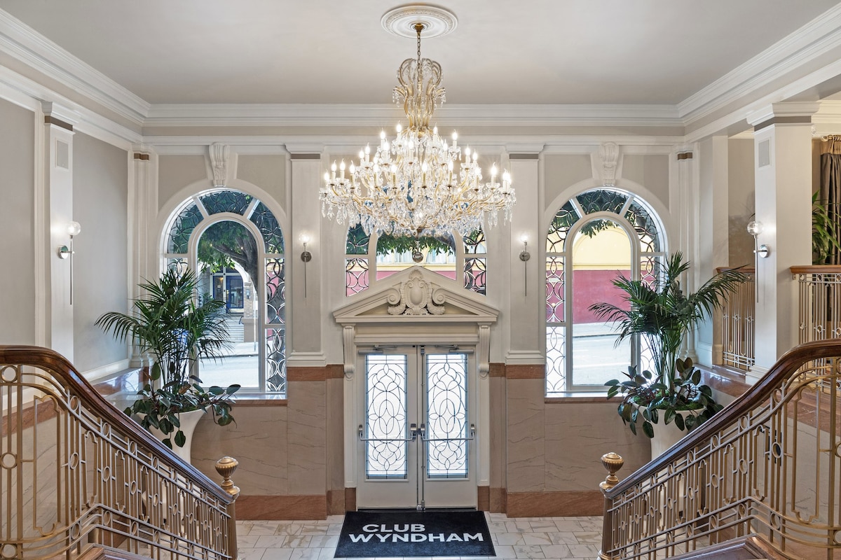 A grand entrance is highlighted by a sparkling chandelier that hangs from a high ceiling. Elegant arched windows allow natural light to illuminate the lobby, where decorative plants are positioned in the corners. A welcome mat displays the name of the resort, enhancing the inviting atmosphere.