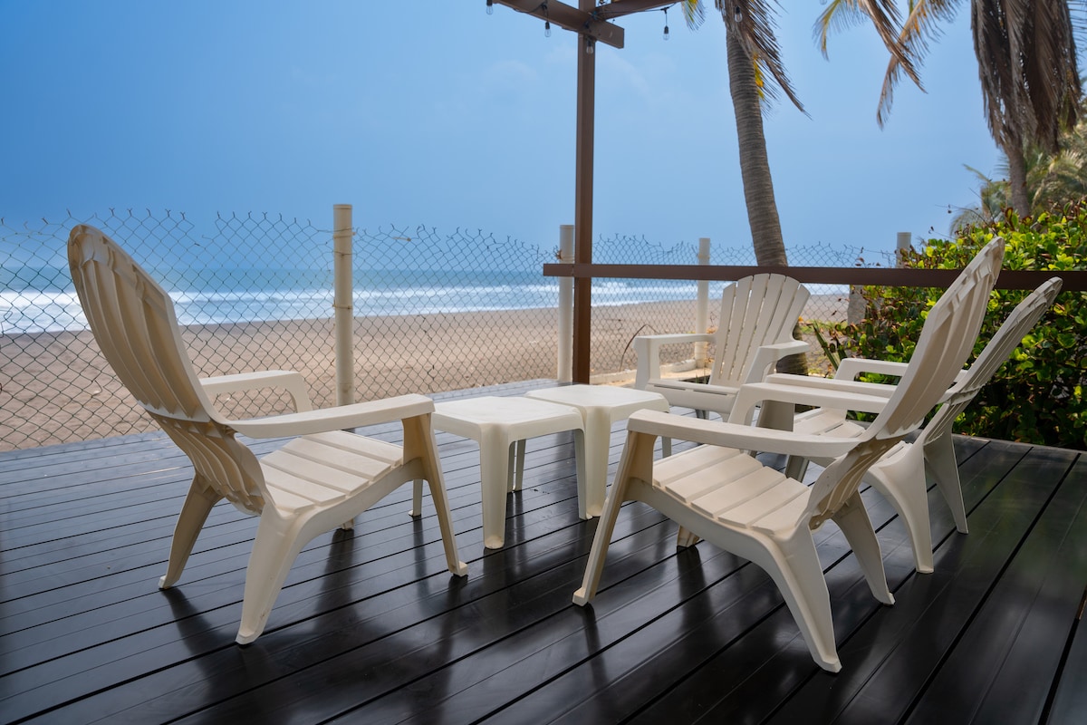 A patio space is highlighted by four white lounge chairs arranged around a small table. The wooden deck offers a view of the sandy beach and ocean in the distance, framed by palm trees and a protective fence.